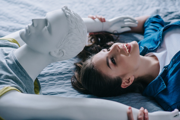 Zijaanzicht van de lachende vrouw en mannequin liggend op bed, eenzaamheid concept - Foto, afbeelding