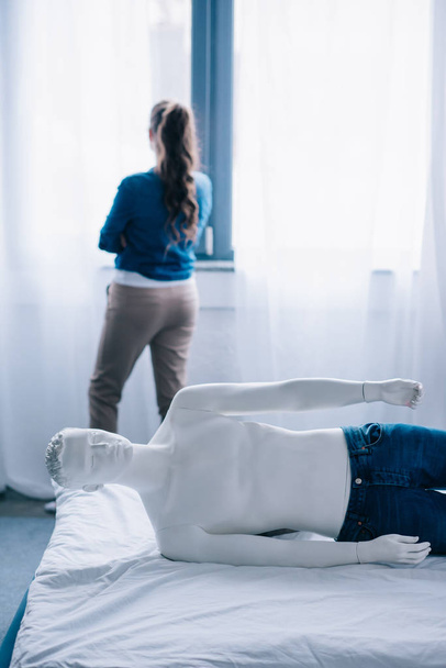 Achteraanzicht van de vrouw stond op venster en leek pop liggend op bed, eenzaamheid concept - Foto, afbeelding