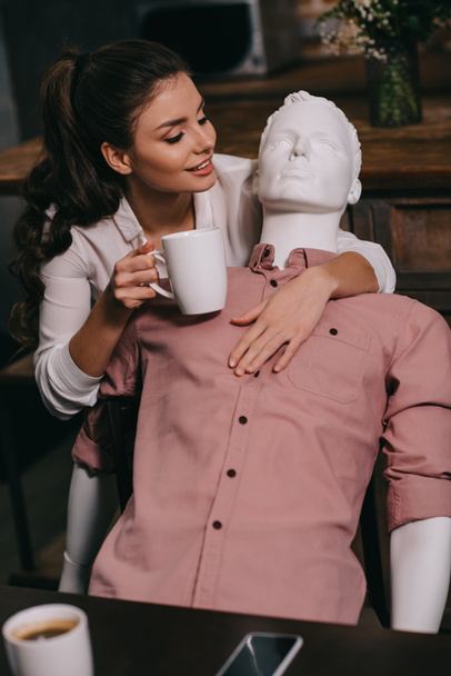 jovem com xícara de café abraçando manequim em casa, conceito de sonho de relacionamento perfeito
 - Foto, Imagem