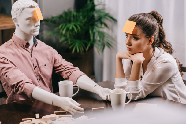 triest jonge vrouw met sticky note op voorhoofd zitten aan tafel met mannequin, eenzaamheid concept - Foto, afbeelding