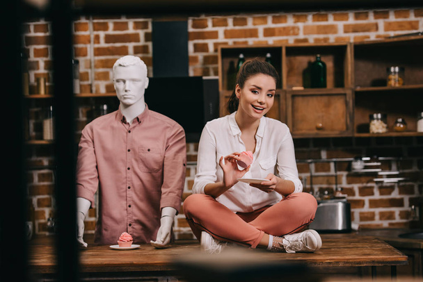 woman with cupcake sitting on table with mannequin behind, loneliness concept - Photo, Image
