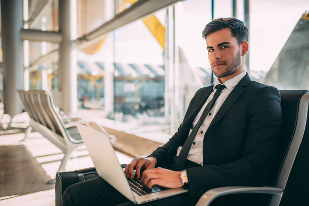 Молодой бизнесмен сидит за компьютером с чемоданом в аэропорту в ожидании рейса
 - Фото, изображение