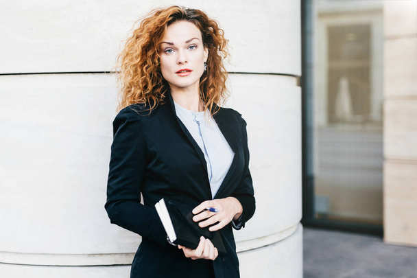 Young beautiful woman wearing formal clothes, dressed formally, holding pocketbook with pen, having bushy hairstyle, standing near office builduing. Successful pretty female office worker working - Photo, Image
