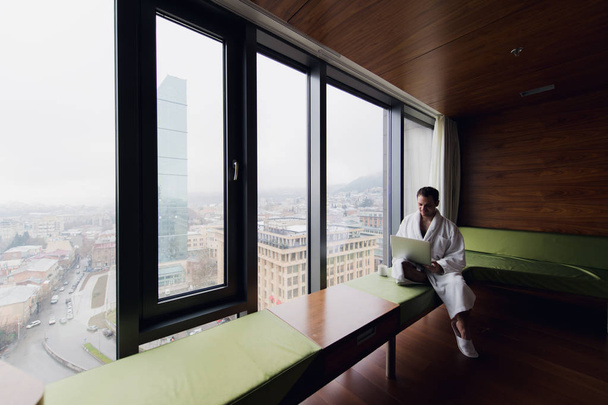 Handsome young businessman wearing bath robe on the bed with laptop in modern room. Self-employed person using laptop in penthouse and looking at sunny city view in window - Photo, Image