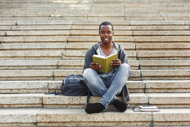 Smiling african-american student reading book on university stairs - Foto, imagen