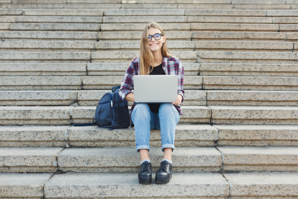 Hymyilevä opiskelija istuu portaissa kannettavan tietokoneen avulla
 - Valokuva, kuva