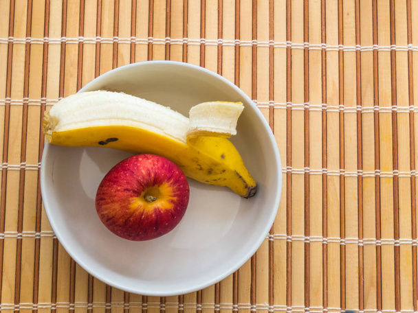 Zdrowe przekąski, banan i jabłko, wewnątrz białe miski na drewnianym stole. - Zdjęcie, obraz