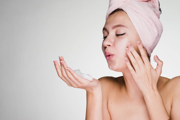 a cute young girl with a pink towel on her head applies a cleansing foam on her face - Fotó, kép