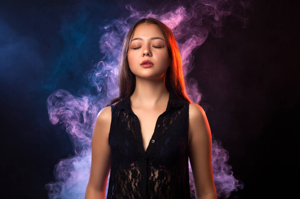 Asian dark-haired woman in a black dress posing against a background of red and red smoke from a vape on a black isolated background - Photo, Image