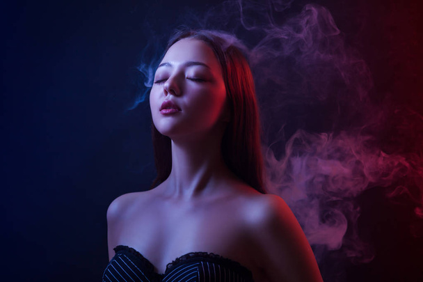 Ázsiai fiatal nő lehunyta a szemét, és pózol a háttér, a piros és a kék füst elszigetelt fekete alapon a vape - Fotó, kép