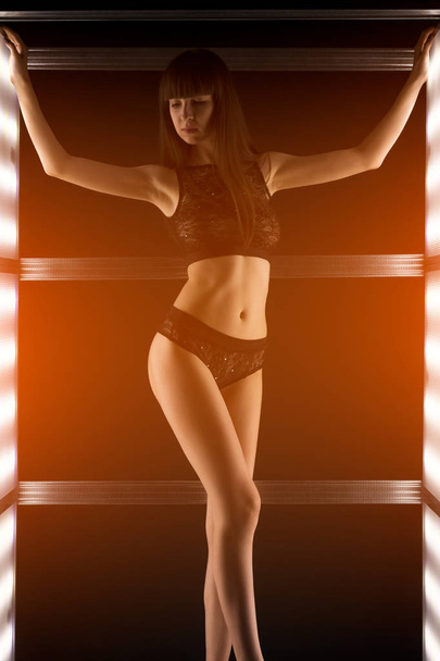 Kaunis seksikäs nainen mustissa alusvaatteissa poseeraa valoseinällä pimeässä studiossa
 - Valokuva, kuva