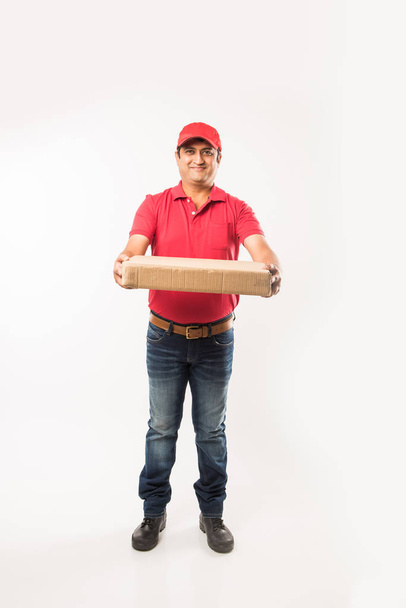 Концепция доставки - портрет счастливого индийца или курьера в красной или зеленой униформе, идущего или прыгающего, проверяющего или представляющего картонную коробку с копировальным местом
 - Фото, изображение