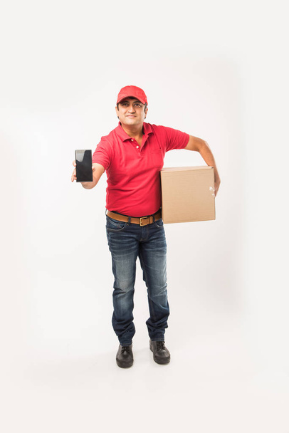 Concept de livraison - Portrait de Happy Indian ou livreur en uniforme rouge ou vert marchant ou sautant ou vérifiant ou présentant boîte en carton avec espace de copie
 - Photo, image