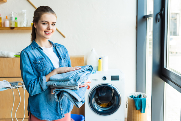 young woman holding clothes and smiling at camera while standing near washing machine at home - Photo, Image
