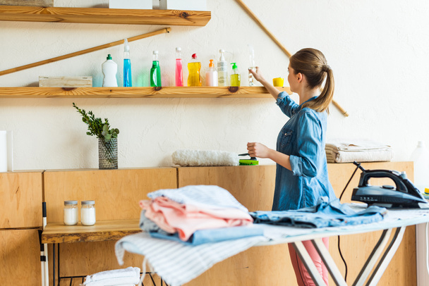 自宅の服をアイロンしながら洗浄液を選択する若い女性 - 写真・画像