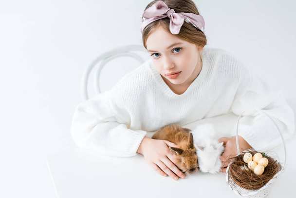 schönes Mädchen blickt in die Kamera, während es mit pelzigen Kaninchen und Korb mit Ostereiern im Nest auf weiß sitzt - Foto, Bild