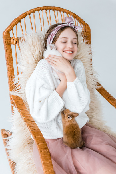 beautiful happy teenage girl hugging rabbits while sitting in rocking chair on white - Zdjęcie, obraz