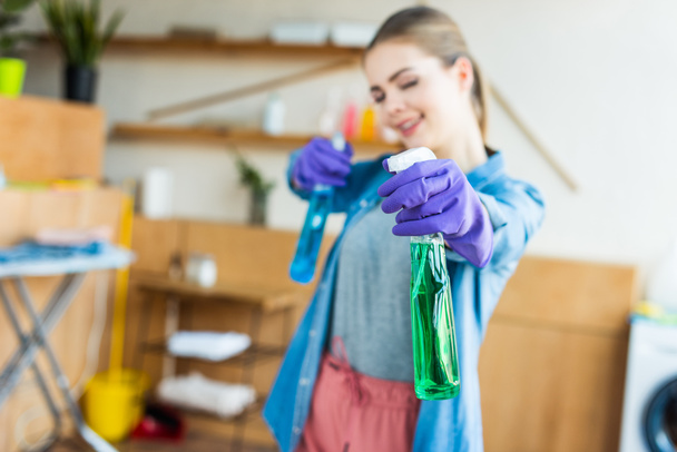 close-up view of smiling young woman in rubber gloves holding spray bottles with cleaning fluids - Foto, Imagem