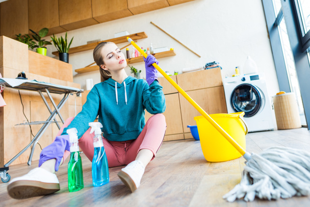 молодая женщина в резиновых перчатках держит швабру и сидит на полу во время уборки дома
 - Фото, изображение