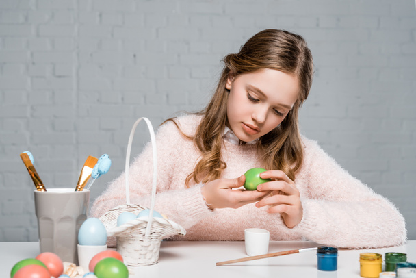 beautiful little girl holding easter egg while sitting at table with pants and brushes - Photo, Image