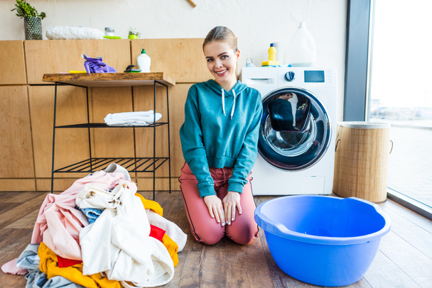 beautiful young woman smiling at camera while kneeling between laundry and plastic basin - Photo, Image