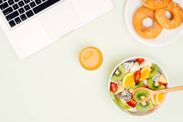 Υγιή επιχειρηματική μεσημεριανό στο γραφείο, φρούτα σαλάτα μπολ κοντά στο λευκό φόντο τον φορητό υπολογιστή. Βιολογικό φαγητό και ντόνατς. Σνακ στα διαλείμματα. - Φωτογραφία, εικόνα