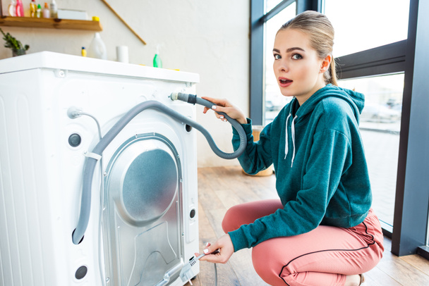 shocked young woman looking at camera while crouching near broken washing machine - Foto, Imagen