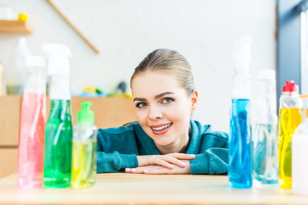 mujer joven sonriendo a la cámara mientras está sentada en la mesa con botellas de plástico con productos de limpieza
 - Foto, imagen