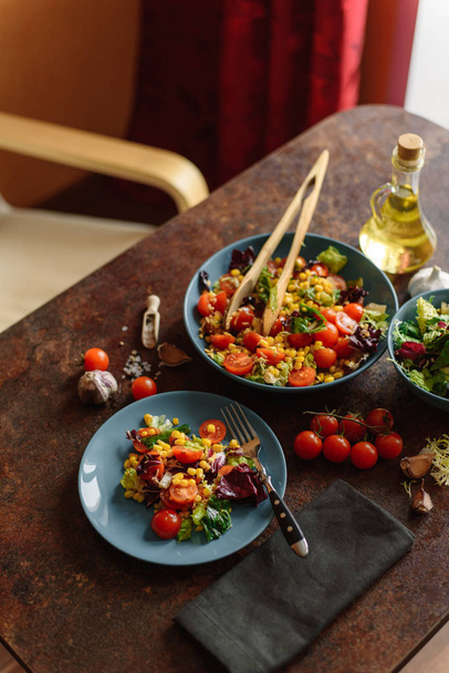 Ensalada vegetariana con maíz, tomates cherry, lechuga, col y perejil con aderezo picante servido en platos azules con tenedor, pinzas, aceite de oliva y servilleta en mesa de madera
. - Foto, imagen