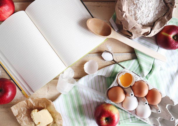 γκρο πλαν του άδειο ανοιχτό βιβλίο με ξύλινο κουτάλι, κόκκινα μήλα, βούτυρο και τα αυγά σε φόντο ξύλινο τραπέζι στην κουζίνα. ψήσιμο έννοια - Φωτογραφία, εικόνα