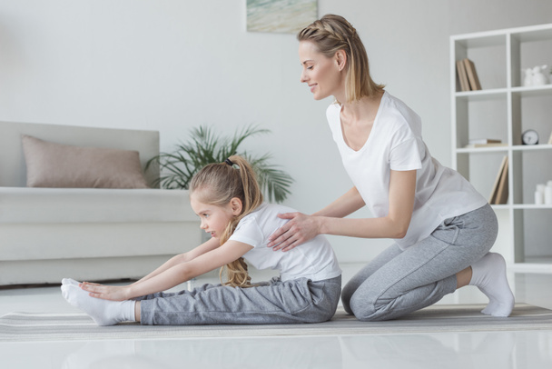 mother helping her daughter to do forward bend at home - Photo, Image
