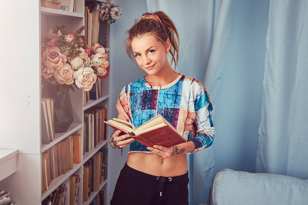 Joven chica tatuada de pie cerca de una estantería leer un libro en casa
. - Foto, imagen