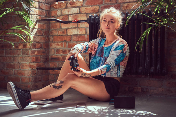 Uma menina tatuada beleza em roupas casuais senta-se no chão e joga jogos de vídeo em uma sala com interior loft contra uma parede de tijolo
. - Foto, Imagem