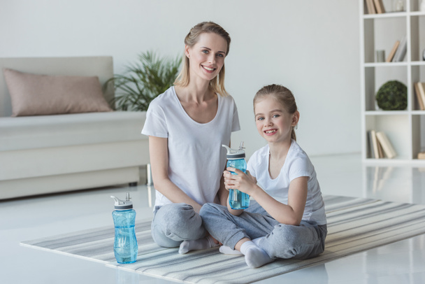 Anne ve kızı ile fitness su şişeleri yoga paspaslar üzerine oturan - Fotoğraf, Görsel