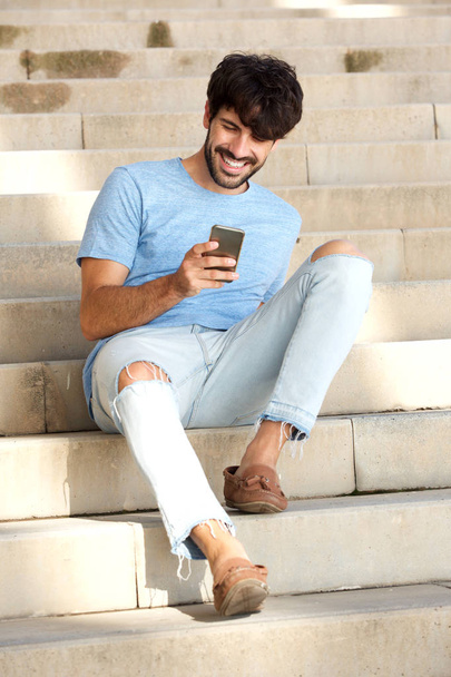 Portrait of friendly young man sitting on steps looking at mobile phone - Photo, Image