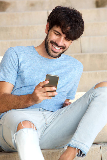 Πορτρέτο του χαρούμενος νεαρός άνδρας κάθεται στο βήματα με smartphone - Φωτογραφία, εικόνα