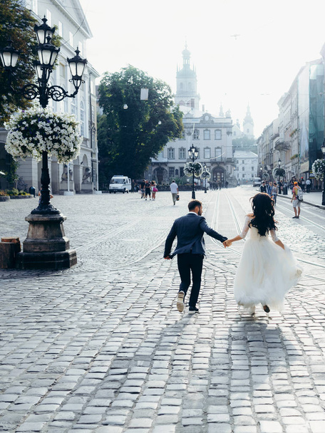 Cheerful beautiful newlywed couple is holding hands while running along the sunny town street. Vertical back view. - Fotó, kép
