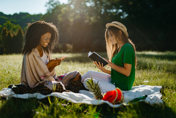 Piknik dwójki przyjaciół dziewczyna wielu wyścigu. Piękne dziewczyny Blondynka jest czytanie książki, podczas gdy jej szczęśliwy uśmiechający się przyjaciel Afryki jest na czacie i przeglądania za pomocą telefonu komórkowego. - Zdjęcie, obraz
