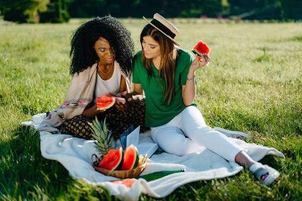 zwei multiethnisch schöne lächelnde Freundinnen chatten und surfen über das Handy, während sie beim Picknick die Wassermelone essen. - Foto, Bild