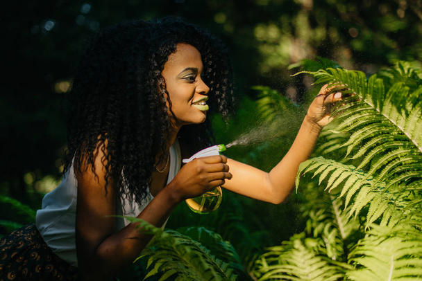 Side outdoor portrait of the attractive young african girl with green lipstick is using the green plastic trigger spray for ferns. - Foto, immagini