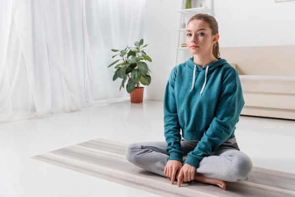 girl sitting in lotus position on yoga mat and looking at camera at home - Foto, afbeelding