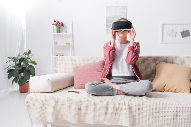 Mädchen sitzt in Lotusposition mit Virtual-Reality-Headset auf dem heimischen Sofa - Foto, Bild
