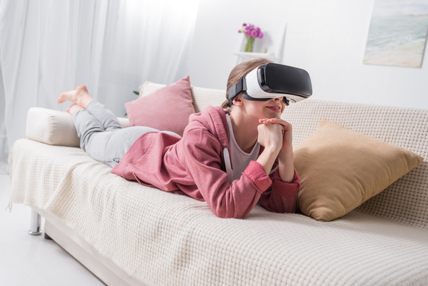 glückliches Mädchen, das auf dem Sofa liegt und zu Hause etwas mit Virtual-Reality-Headset anschaut - Foto, Bild