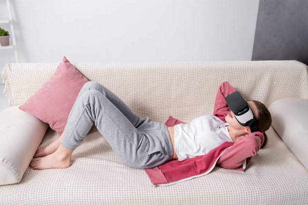high angle view of girl doing sits up with virtual reality headset on sofa at home - Photo, Image