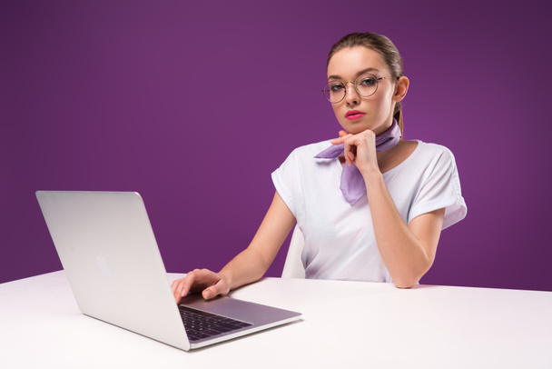 pensive girl using laptop and looking at camera isolated on purple - Φωτογραφία, εικόνα