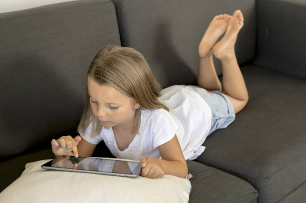young sweet and happy little girl 6 or 7 years old lying on home living room sofa couch using internet digital tablet touch pad  - Photo, Image
