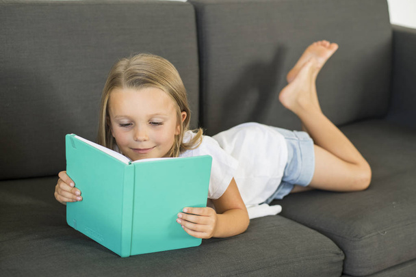 young sweet and happy little girl 6 or 7 years old lying on home living room sofa couch reading a book quiet and adorable in children education - 写真・画像