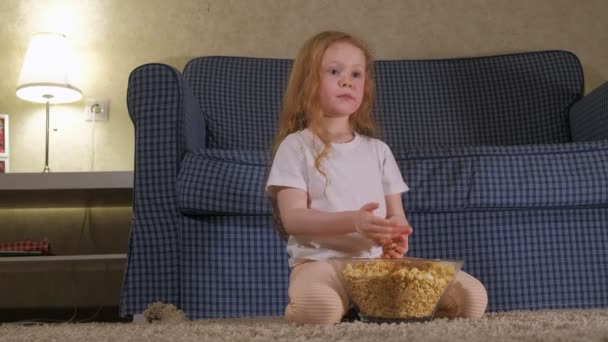 A little girl is sitting on the couch watching TV and eating popcorn - Séquence, vidéo