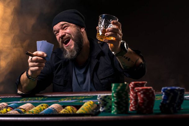 Hombre barbudo con cigarro y vidrio sentado en la mesa de póquer y gritando aislado en negro
 - Foto, Imagen