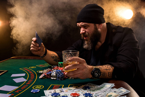 Brodaty mężczyzna z cygara i szkła siedzi przy stole w kasynie. Hazard, gry w karty i ruletka. - Zdjęcie, obraz